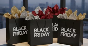 Three black Black Friday shopping bags filled with various insurance documents and policy paperwork, arranged on a clean wooden desk with a pen, tablet, and a notebook. In the background, a modern office window overlooks a city skyline at dusk.