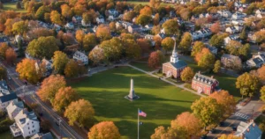 Aerial view of the Lexington Battle Green in autumn, showing the central green, a tall monument, a flagpole with the American flag, and historical red-brick buildings surrounded by homes and trees in peak fall color.